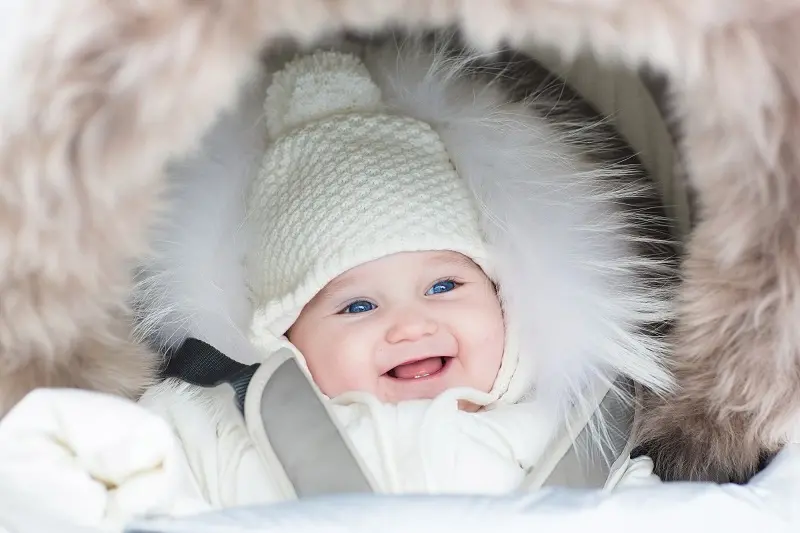 Happy smiling baby wearing a white knit ha and white winter coat in a stroller.