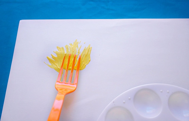 Orange plastic fork rubbing yellow paint onto white paper.