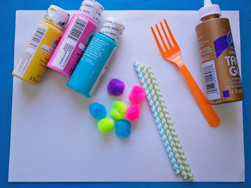 Acrylic paints, pom poms, chevron paper straws, plastic fork, and tacky glue laying on paper.