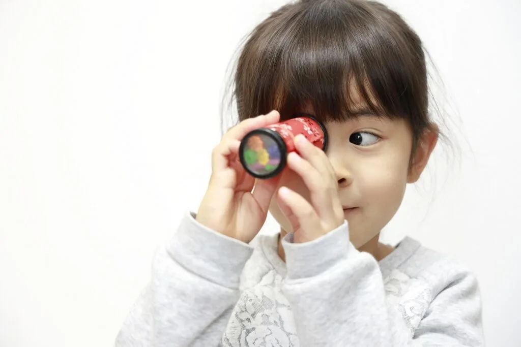 Young Japanese girl wearing a white long sleeved shirt holding a toy kaleidoscope up to her eye.