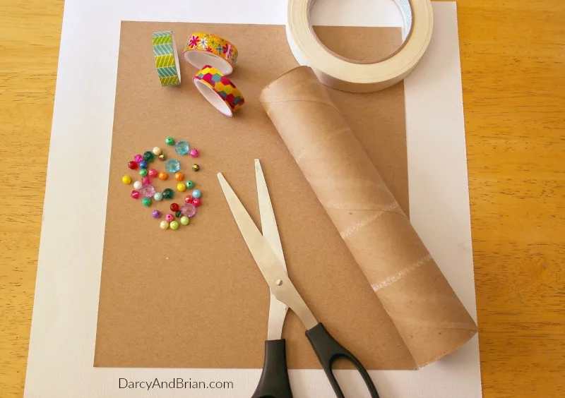 Craft supplies needed to make your own homemade kaleidoscope for kids. Includes scrapbooking paper, rolls of washi tape, empty paper towel roll, scissors laying on table.