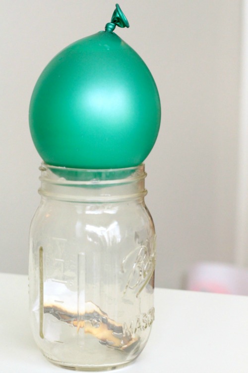 Green balloon sitting on top of a glass jar. A piece of paper is on fire inside the jar.