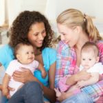 Two moms sitting on the couch with babies talking and laughing.