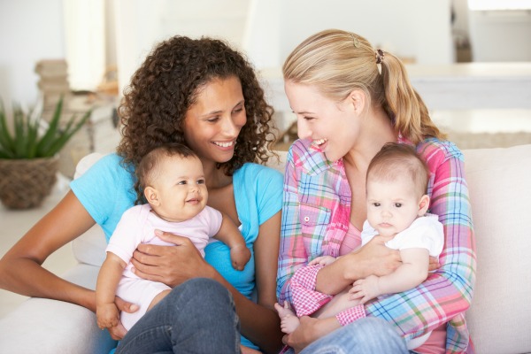 Two moms sitting on the couch with babies talking and laughing.