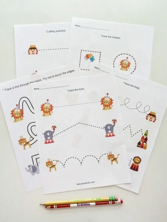 Several pages printed out from the preschool circus printable pack. The pages are overlapping on a white desk. A writing pencil and a colored pencil are lined up along the bottom.