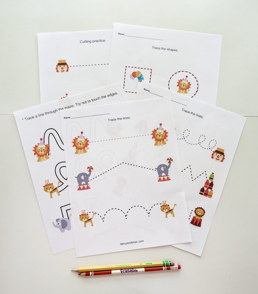 Several pages printed out from the preschool circus printable pack. The pages are overlapping on a white desk. A writing pencil and a colored pencil are lined up along the bottom.