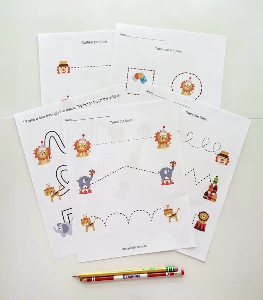 Several pages printed out from the preschool circus printable pack. The pages are overlapping on a white desk. A writing pencil and a colored pencil are lined up along the bottom.