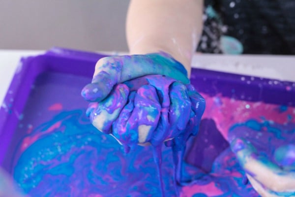 Child's hand holding blue, purple, and pink oobleck in their fist as it oozes out. A tray beneath their hand has more.