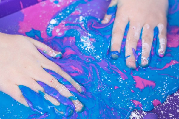 A pair of white hands laying in galaxy colored oobleck.