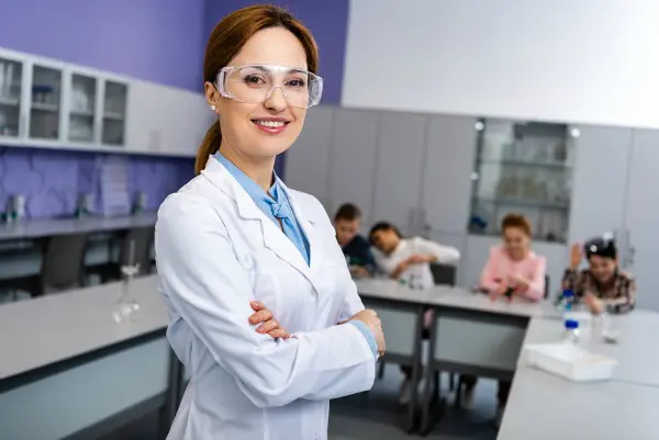 Female scientist wearing lab coat and goggles in classroom science lab setting with students
