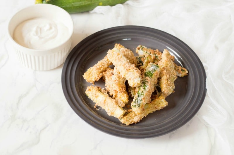 Finished baked zucchini fries on black plate with small white bowl with dipping sauce