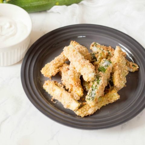 Baked zucchini fries piled on black plate with dip in small white ramekin