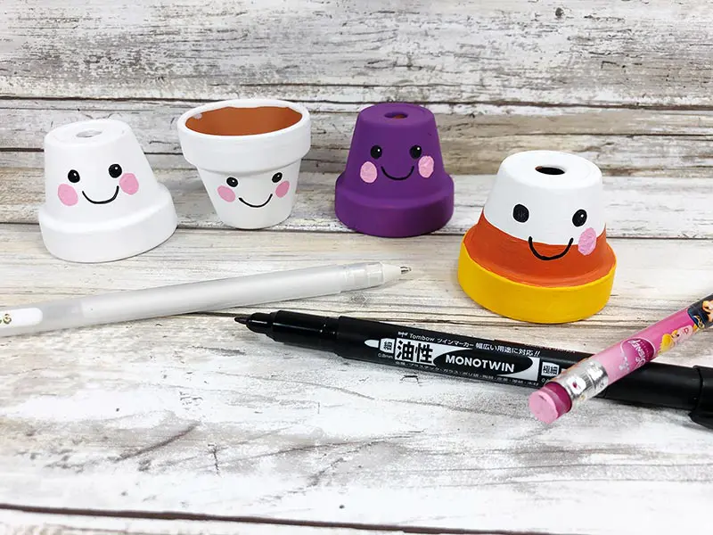 Four little flower pots painted to look like ghosts, candy corn, and spider on a white board background. Showing how to add a cute Kawaii style face to the clay pots.