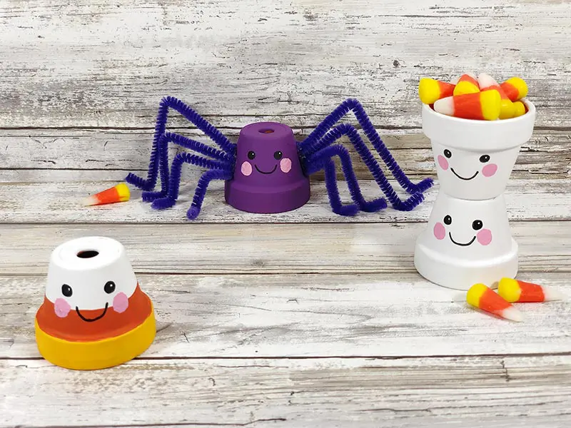 Finished Kawaii Halloween flower pots. One painted in candy corn colors, a purple spider, and two ghosts stacked on top of each other. All have cute Kawaii inspired faces.