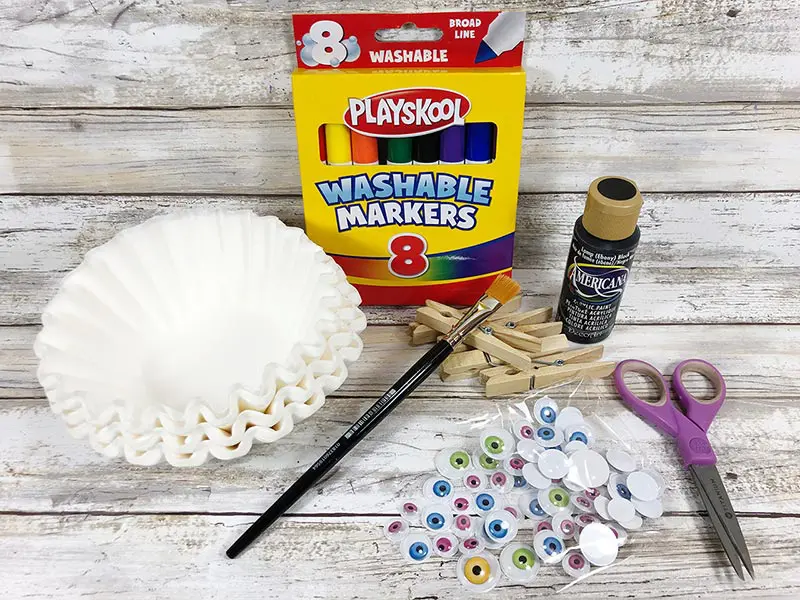 Stack of white coffee filters, box of markers, googly eyes, black craft paint, clothespins, paint brush, and scissors set out for project