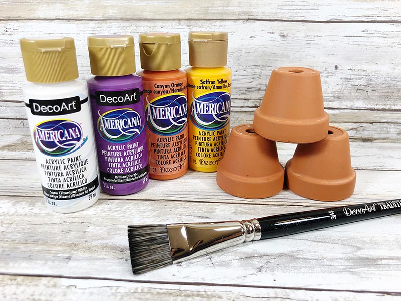 Bottles of white, purple, orange, and yellow acrylic craft paint next to three mini flower pots and a paintbrush.