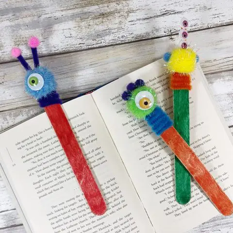 Three craft stick monster bookmarks laying on open book. The bookmarks are multiple colors.