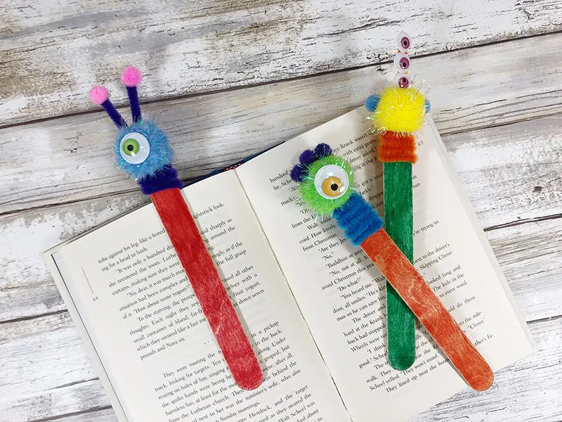 Three craft stick monster bookmarks laying on open book. The bookmarks are multiple colors.