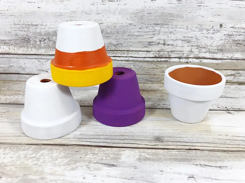 Four mini clay pots painted without faces added. Two are painted white, one purple, one with white, orange, and yellow.