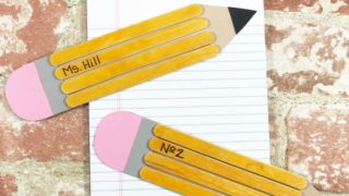Two school pencils made with paper and wooden craft sticks on notebook with brick background