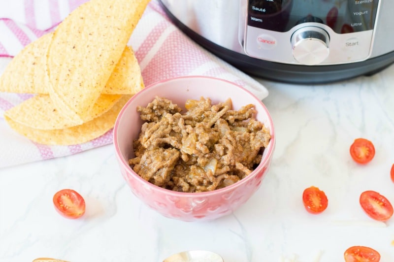 Cooked taco meat in pink bowl on counter by taco shells, tomatoes, and Instant Pot