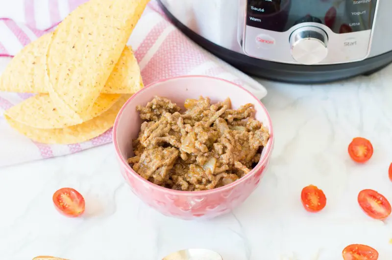 Cooked taco meat in pink bowl on counter by taco shells, tomatoes, and Instant Pot