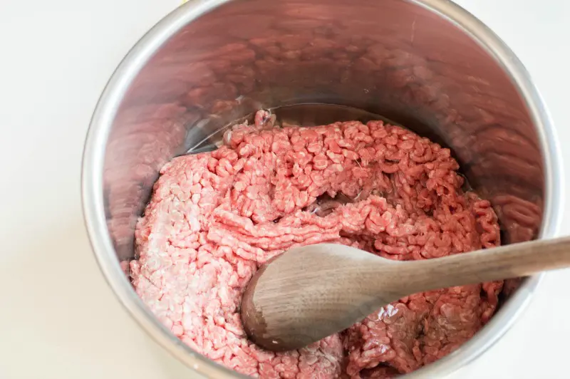 Wooden spoon breaking up fresh ground beef in Instant Pot inner pot