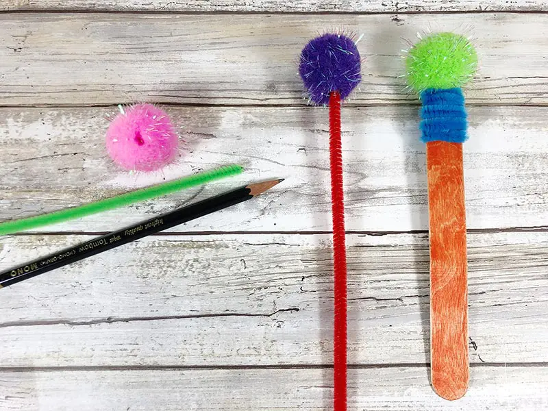 Pink pom pom and green pipe cleaner laying on white board background. Showing steps to assemble monster bookmark.
