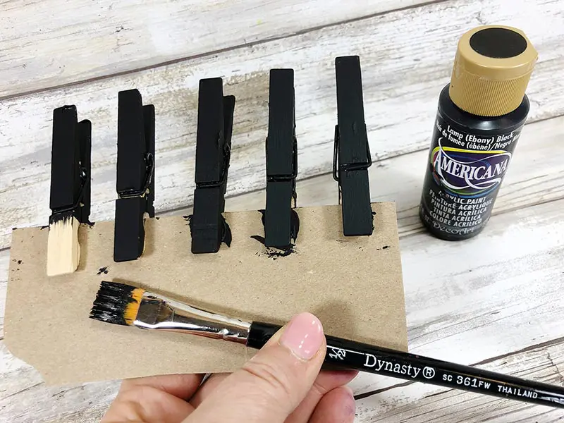 Five clothespins clipped on piece of cardboard being painted black with paintbrush