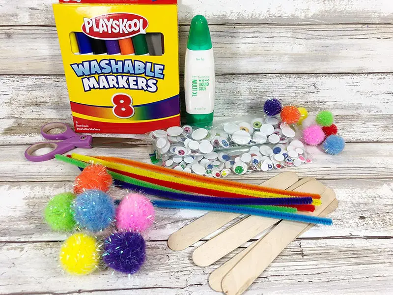 Box of markers, glue, googly eyes, assorted pom poms, craft sticks, and chenille stems laying on white board background.