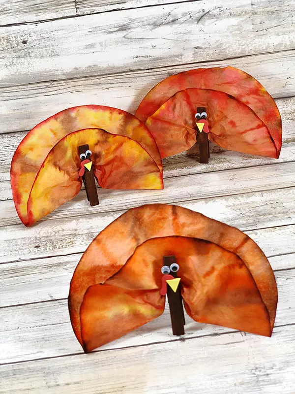 Three finished turkeys made out of coffee filters and clothes pins standing on white wood background.