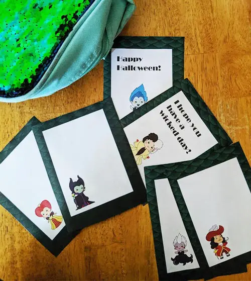 Printed Disney Villain lunch note cards cut apart and spread out on wood table with green and blue lunch box nearby.