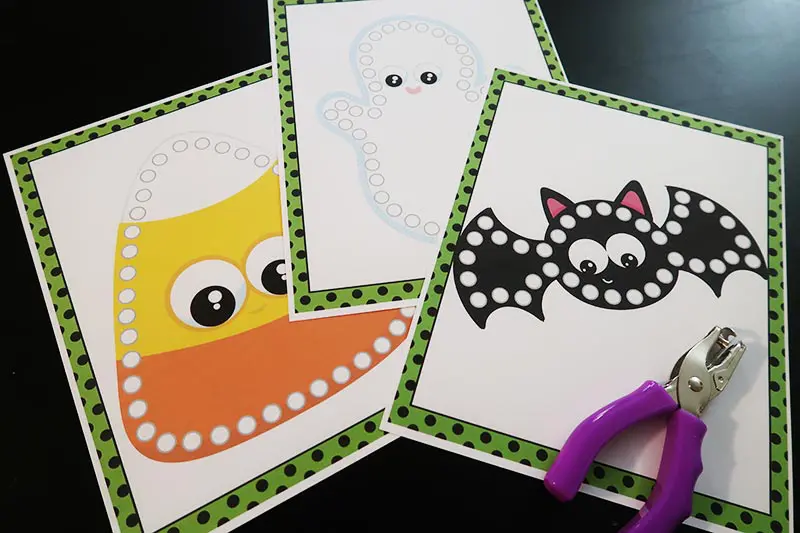 Candy corn, ghost, and bat lacing cards printed on cardstock paper and laying on table next to hole puncher.