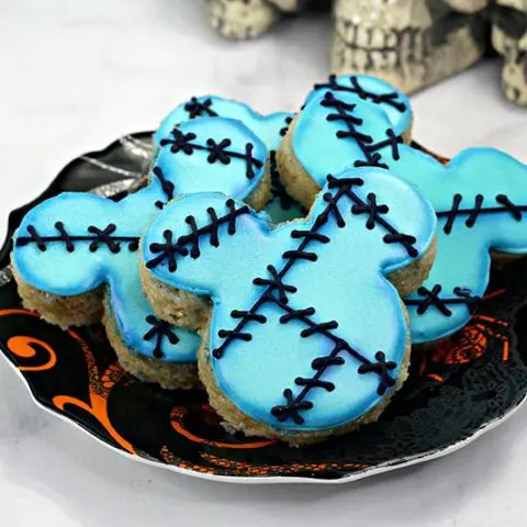 Mickey shaped rice crispy treats decorated with blue icing and black stitch marks. Several arranged on a Halloween plate.