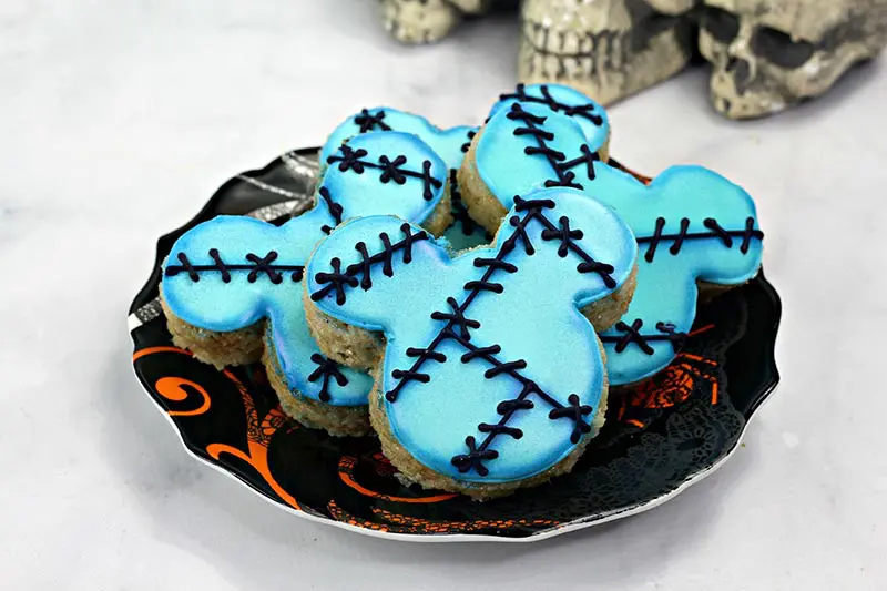 Mickey shaped rice crispy treats decorated with blue icing and black stitch marks. Several arranged on a Halloween plate.