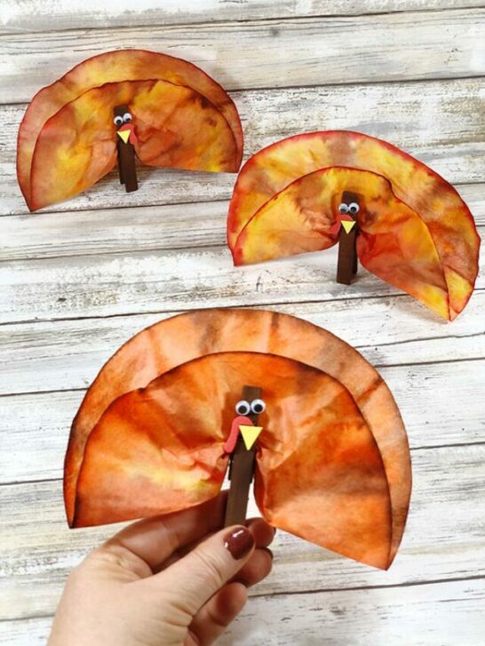 Three completed coffee filter turkey craft projects. Woman's hand holding one turkey. Two turkeys standing on white wood background.