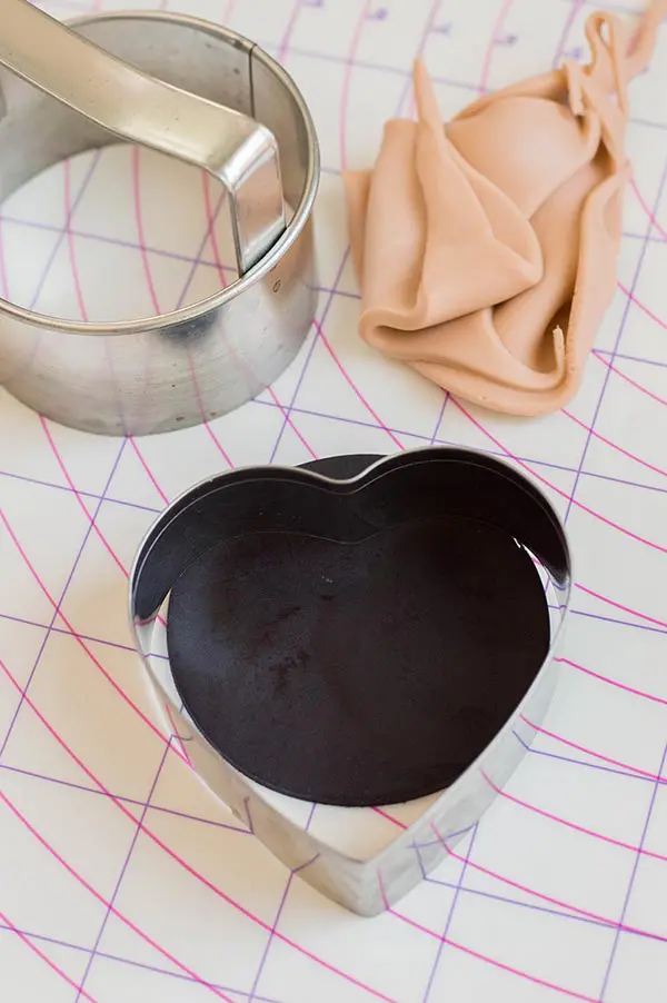 Heart shaped cookie cutter on black fondant. Circle cookie cutter and beige fondant scraps on pastry mat.