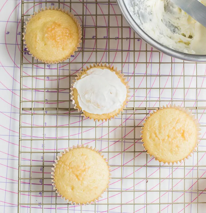 Thinly frosting cupcakes. Cupcakes are on wire cooling rack next to bowl of frosting.