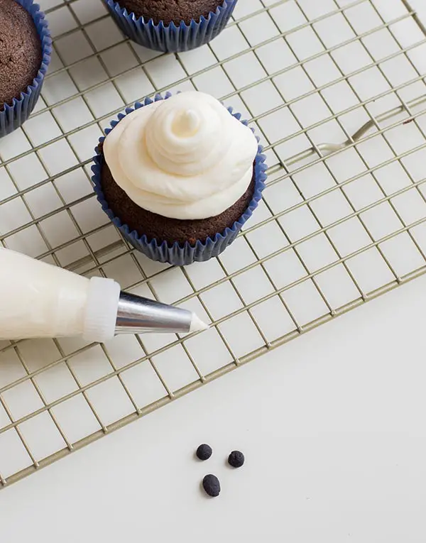 Frosted chocolate cupcake and piping bag on cooling rack. Black fondant rolled in three small balls on white countertop.