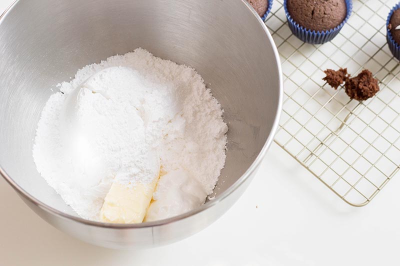 Butter, sugar, marshmallow fluff in silver mixing bowl next to cooling rack with chocolate cupcakes