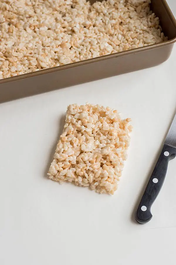 Rectangular piece of rice crispy treat laying on white counter next to knife and pan of cereal treats.