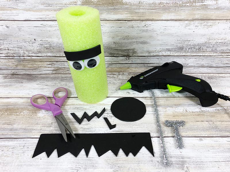 Green pool noodle with googly eyes and black eyebrow attached. DIY decoration in progress.
