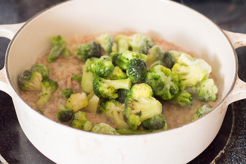 Frozen broccoli florets, tuna, rice, and broth in white cooking pot on stove