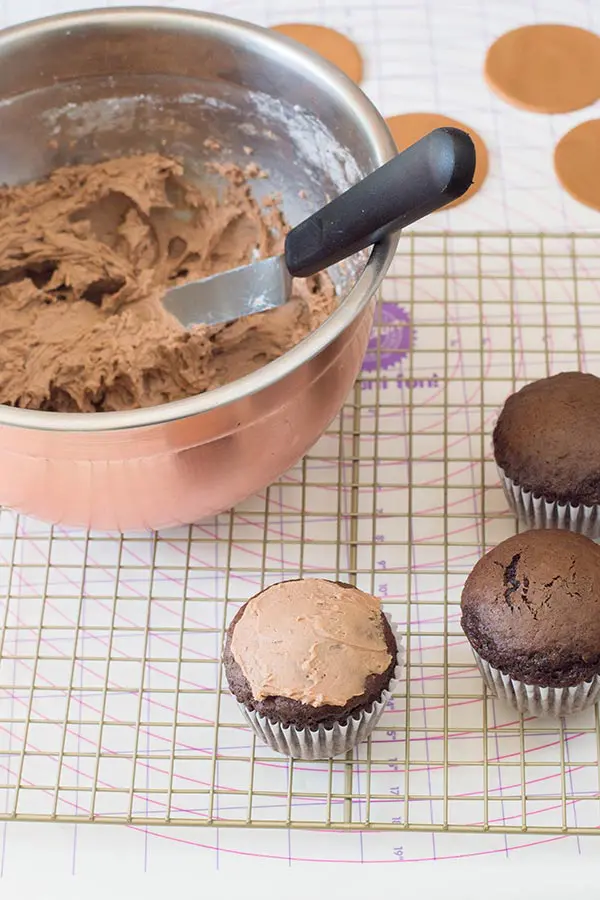 Chocolate buttercream in bowl with offset spatula. One cupcake frosted and two unfrosted cupcakes on wire rack.