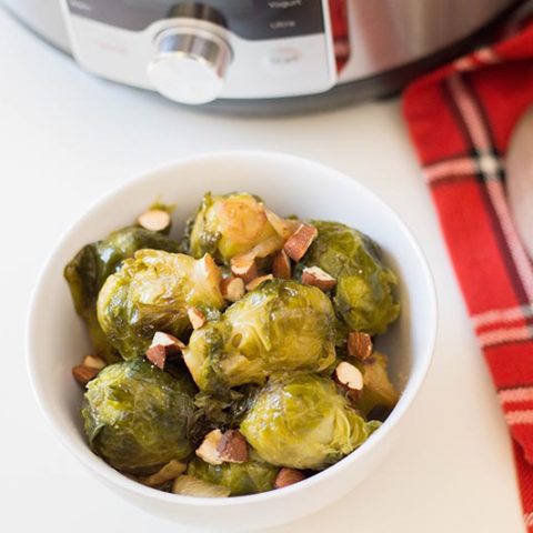 Small white bowl with cooked Brussels sprouts and chopped almonds next to wooden spoon on a red plaid cloth.