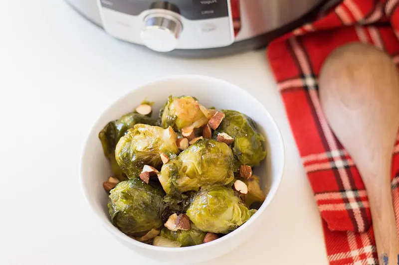 Small white bowl with cooked Brussels sprouts and chopped almonds next to wooden spoon on a red plaid cloth.