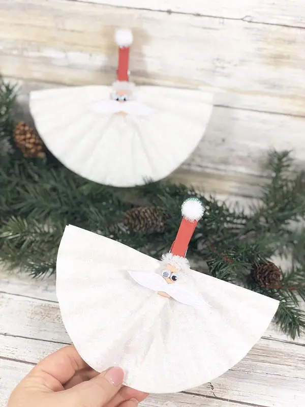 Hand holding one completed coffee filter santa craft and another sitting on evergreen sprig.