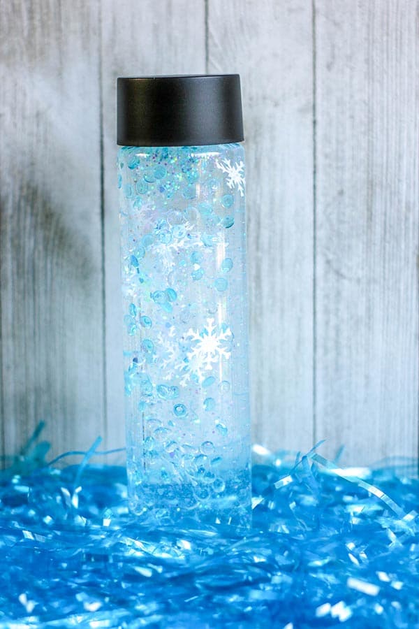 Closed snowflake sensory bottle standing up right on top of blue tinsel.