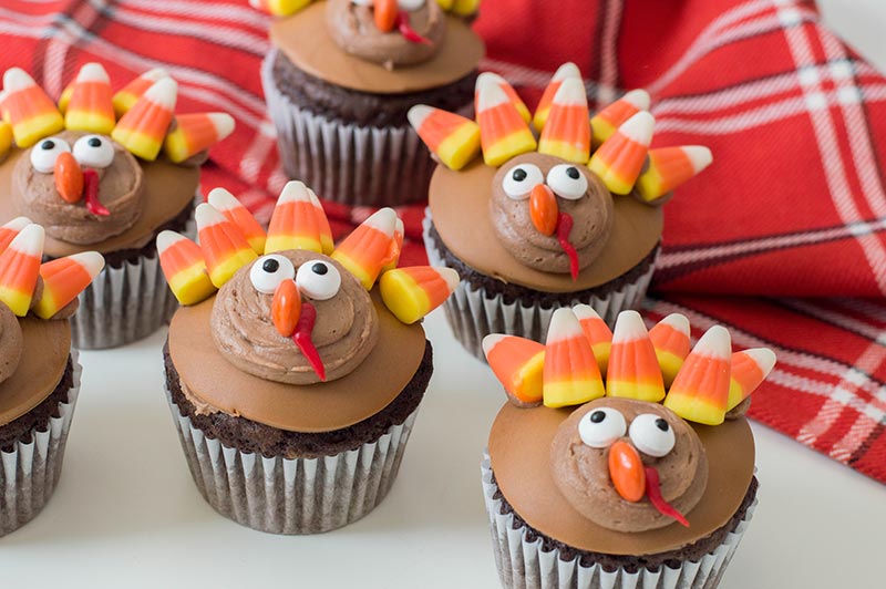Several turkey cupcakes on red plaid cloth and white counter.