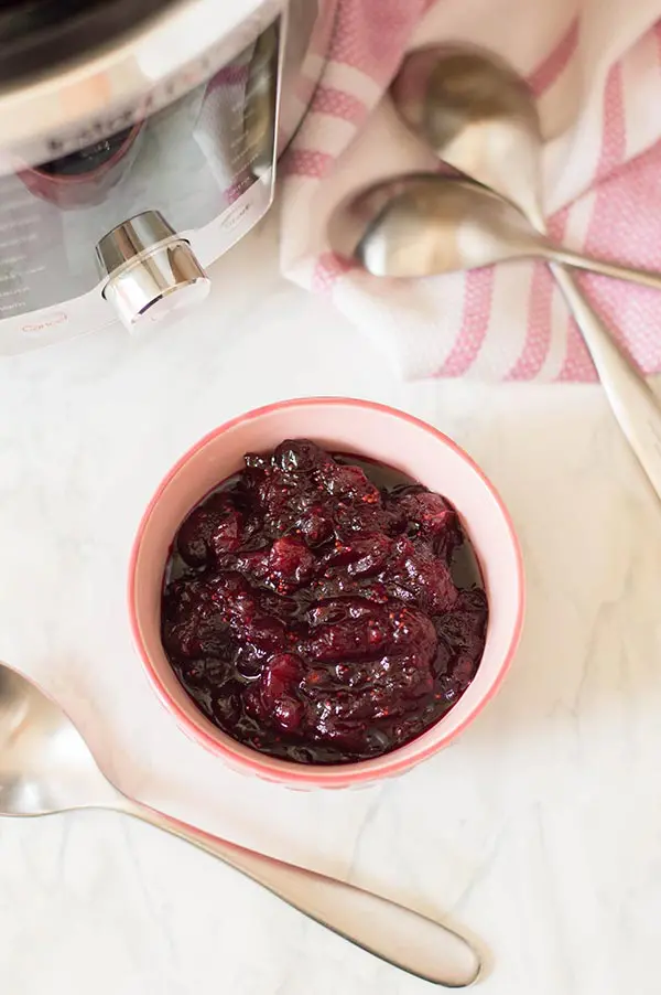 Chunky homemade cranberry sauce in a light pink bowl on counter next to pressure cooker and spoons.